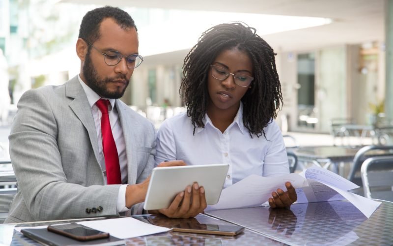 Focused coworkers checking reports on papers and on tablet. Business man and woman sitting in coffee shop, showing tablet and smartphone screens to each other. Teamwork concept
